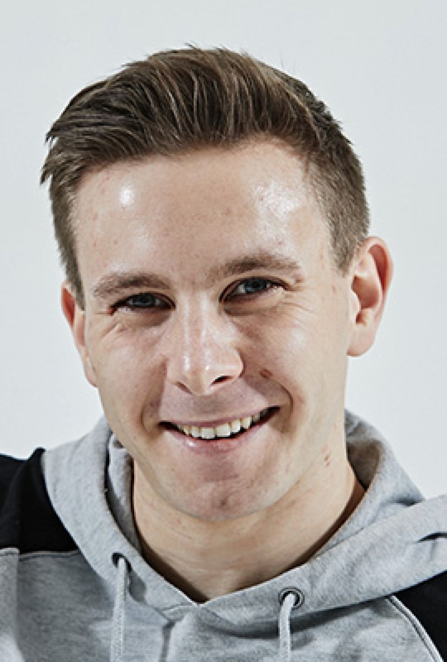 Das Bild zeigt Amber Chiara Eul, mit blondbraunen Haaren, die zu einem Dutt hochgesteckt sind. Sie hat blaue Augen und blickt mit ernstem Ausdruck direkt in die Kamera. Sie trägt einen rosafarbenen Pullover mit einem roten Streifen im Brustbereich. Der Hintergrund ist einfarbig rostbraun.