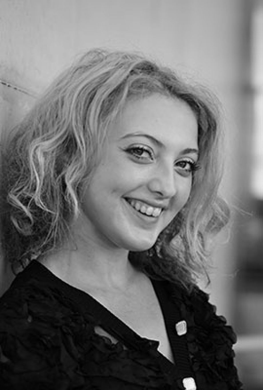 Das Bild zeigt Amber Chiara Eul, mit blondbraunen Haaren, die zu einem Dutt hochgesteckt sind. Sie hat blaue Augen und blickt mit ernstem Ausdruck direkt in die Kamera. Sie trägt einen rosafarbenen Pullover mit einem roten Streifen im Brustbereich. Der Hintergrund ist einfarbig rostbraun.