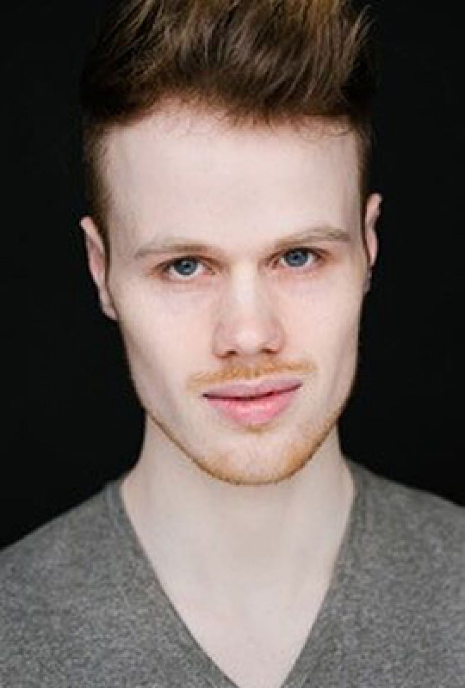 Das Bild zeigt Amber Chiara Eul, mit blondbraunen Haaren, die zu einem Dutt hochgesteckt sind. Sie hat blaue Augen und blickt mit ernstem Ausdruck direkt in die Kamera. Sie trägt einen rosafarbenen Pullover mit einem roten Streifen im Brustbereich. Der Hintergrund ist einfarbig rostbraun.