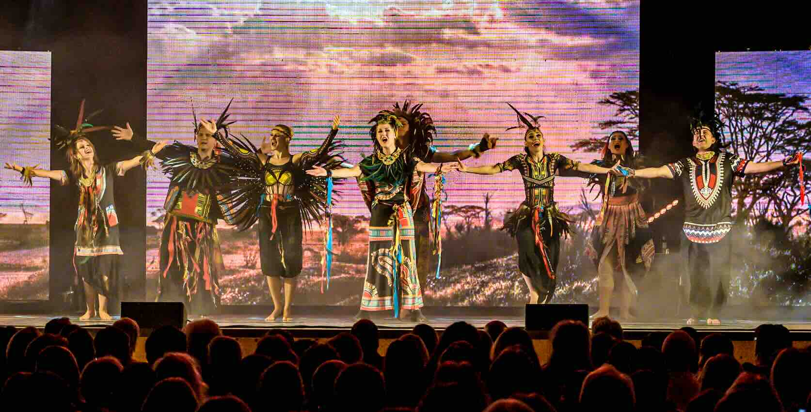Das Bild zeigt eine Szene aus Der König der Löwen: Darsteller in afrikanisch inspirierten Kostümen performen vor einer LED-Wand mit Savannenlandschaft, während das Publikum gebannt zuschaut.
