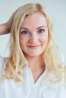 Das Portrait zeigt Verena Mackenberg mit langen, blonden Haaren und einem freundlichen Lächeln. Sie trägt eine weiße Bluse und ihre Hand fasst sich am Hinterkopf ins Haar.