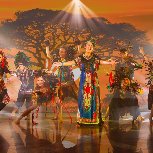 Farbenfrohe Szene aus Der König der Löwen: Tänzer in prachtvollen, afrikanisch inspirierten Kostümen voller Federn und Muster erwecken die Savanne zum Leben. Im Hintergrund ein majestätischer Akazienbaum bei Sonnenuntergang – pure Magie und Lebensfreude!