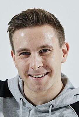 Das Bild zeigt Amber Chiara Eul, mit blondbraunen Haaren, die zu einem Dutt hochgesteckt sind. Sie hat blaue Augen und blickt mit ernstem Ausdruck direkt in die Kamera. Sie trägt einen rosafarbenen Pullover mit einem roten Streifen im Brustbereich. Der Hintergrund ist einfarbig rostbraun.