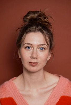 Das Bild zeigt Amber Chiara Eul, mit blondbraunen Haaren, die zu einem Dutt hochgesteckt sind. Sie hat blaue Augen und blickt mit ernstem Ausdruck direkt in die Kamera. Sie trägt einen rosafarbenen Pullover mit einem roten Streifen im Brustbereich. Der Hintergrund ist einfarbig rostbraun.