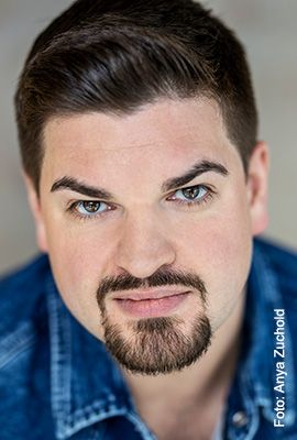 Das Bild zeigt Amber Chiara Eul, mit blondbraunen Haaren, die zu einem Dutt hochgesteckt sind. Sie hat blaue Augen und blickt mit ernstem Ausdruck direkt in die Kamera. Sie trägt einen rosafarbenen Pullover mit einem roten Streifen im Brustbereich. Der Hintergrund ist einfarbig rostbraun.