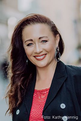 Das Bild zeigt Nadine Hammer, Darstellerin bei Best of Musicals, mit langen, gewellten braunen Haaren und einem freundlichen Lächeln. Sie trägt ein rotes Oberteil, einen schwarzen Blazer und große Ohrringe.