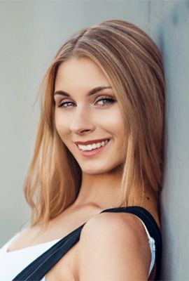 Das Portrait zeigt Nadine Werner mit langen, blonden Haaren. Sie lehnt mit dem Rücken an einer grauen Wand und sieht lächelnd in die Kamera.