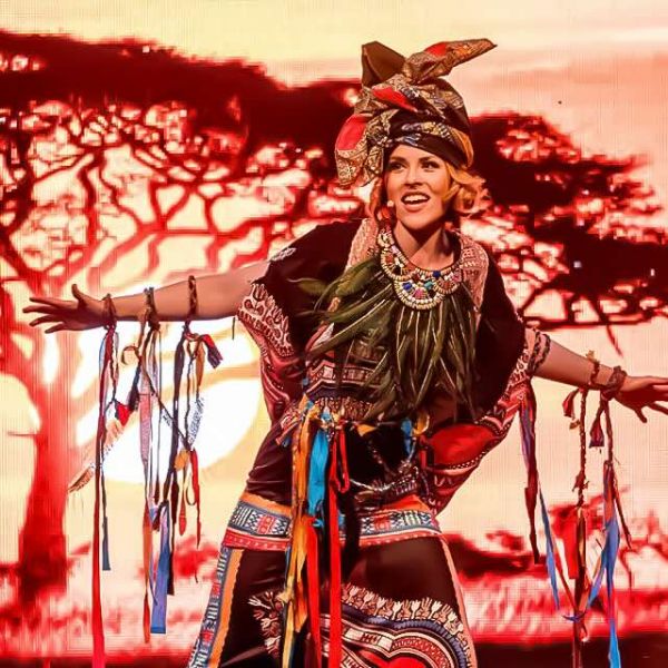 Farbenfrohe Szene aus König der Löwen der Best of Musicals Show: Eine strahlende Darstellerin in afrikanischem Kostüm vor einer eindrucksvollen Savannen-Kulisse!