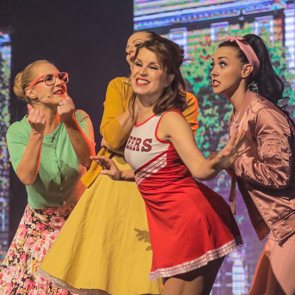 Lebendige Szene aus Grease der Best of Musicals Show: Vier junge Frauen in farbenfrohen Outfits diskutieren leidenschaftlich – mit dabei eine Cheerleaderin und eine Pink Lady.