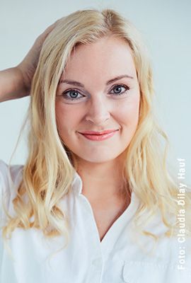 Das Portrait zeigt Verena Mackenberg mit langen, blonden Haaren und einem freundlichen Lächeln. Sie trägt eine weiße Bluse und ihre Hand fasst sich am Hinterkopf ins Haar.