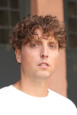 Das Bild zeigt Robert Schmelcher, Darsteller bei Best of Musicals, mit lockigen hellbraunen Haaren und ernsthaftem Blick. Er trägt ein weißes T-Shirt.